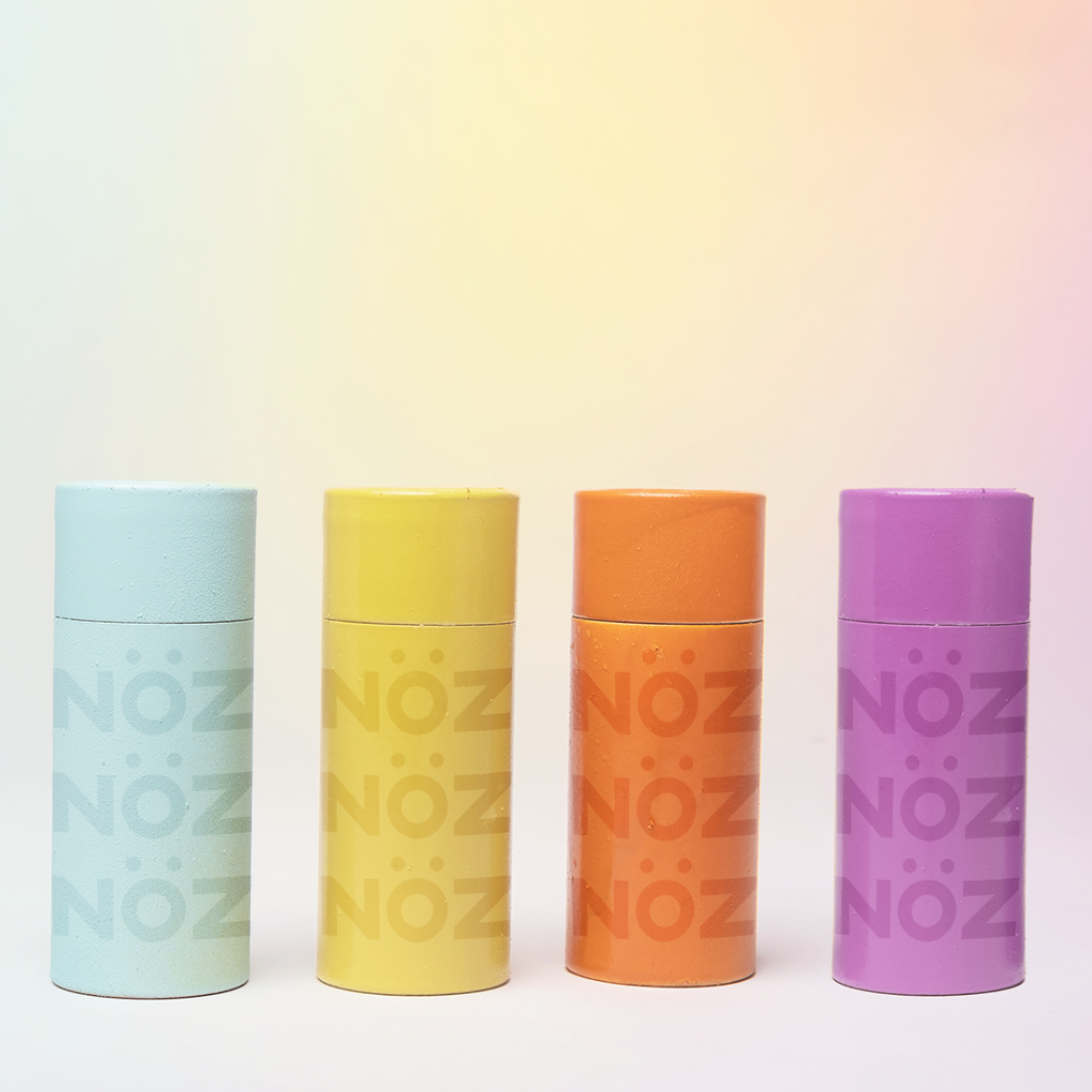 All four Nöz Nözscreen neon color nose sunscreen sticks in blue, yellow, orange, purple products standing next to each other